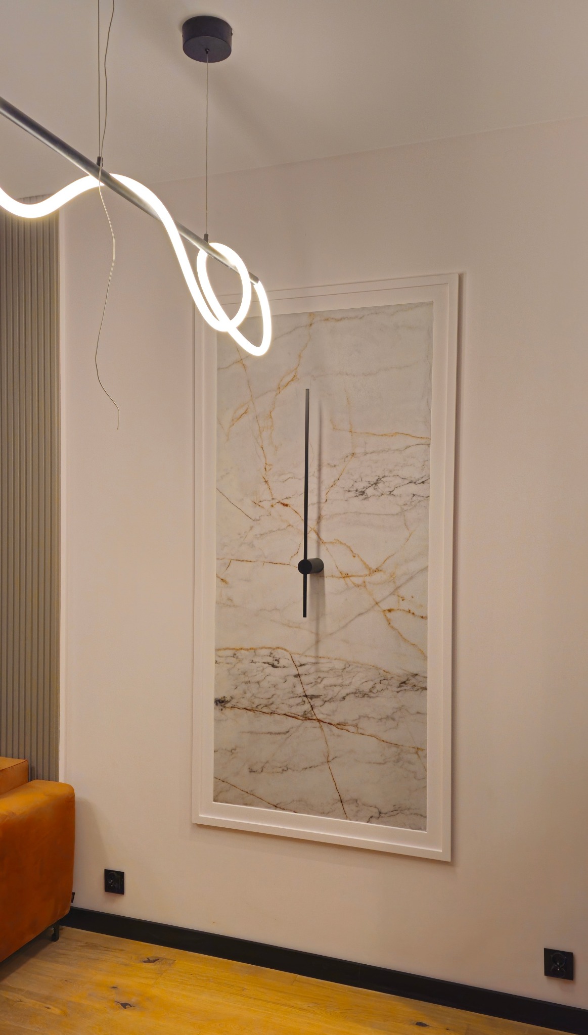 Nowoczesne oświetlenie: abstrakcyjna lampa LED nad obrazem imitującym marmur z minimalistycznym zegarem w salonie. Elementy dekoracyjne i gniazdka elektryczne.
