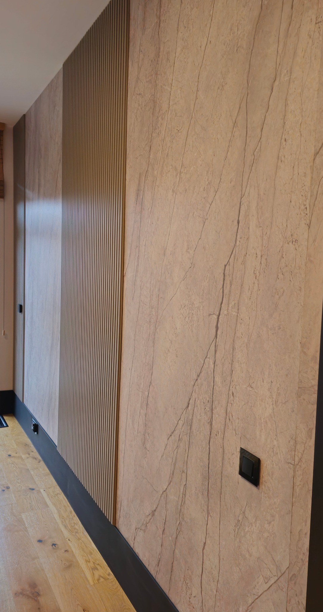 Wykończona ściana z efektem marmuru, panele dekoracyjne z pionowymi listwami i czarna listwa przypodłogowa. Widoczny włącznik światła i fragment drewnianej podłogi.