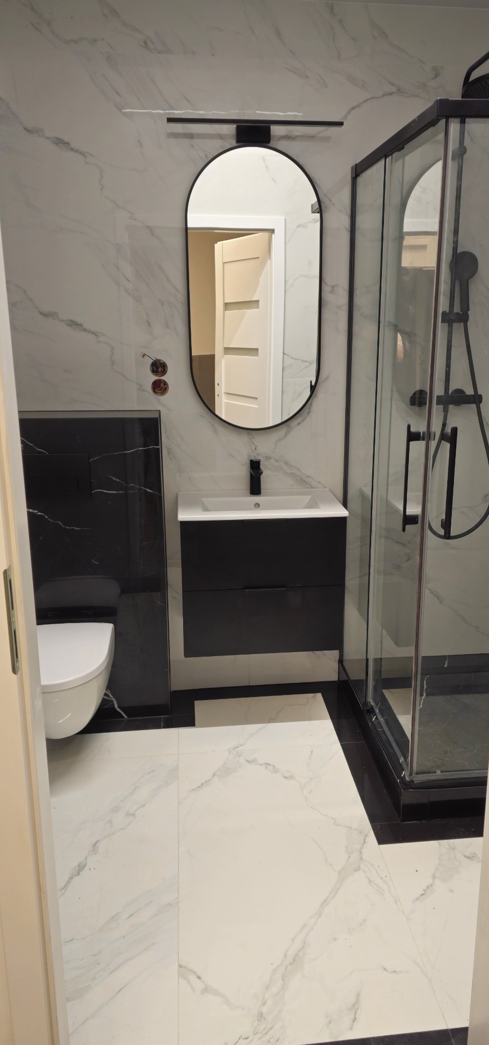 Nowoczesna łazienka z kabiną prysznicową, umywalką z szafką w kolorze czarnym i owalnym lustrem. Białe marmurowe płytki na ścianach i podłodze. Minimalistyczny design.