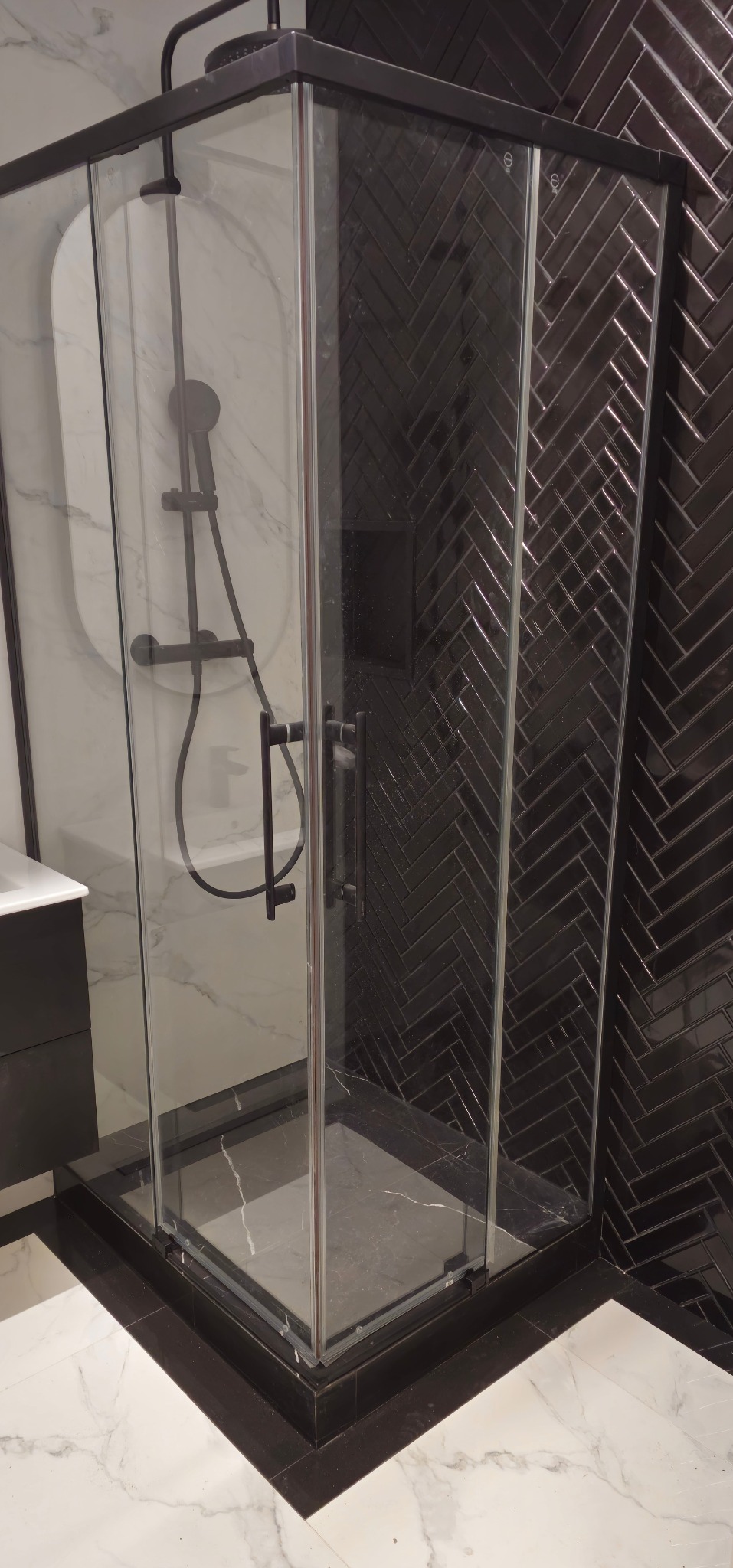 Nowoczesna kabina prysznicowa z czarnymi detalami i armaturą, na tle ściany z ciemnej, błyszczącej glazury ułożonej w jodełkę. Marmurowa posadzka.