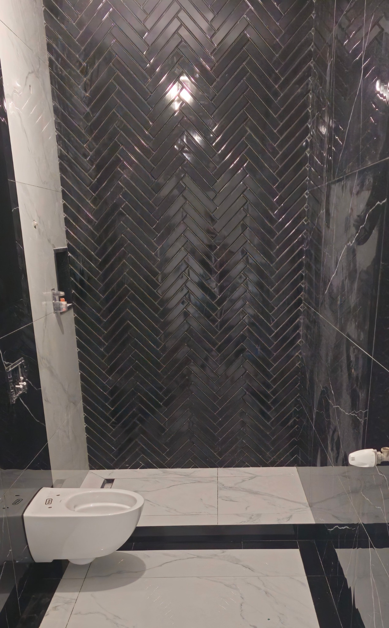 Elegancka łazienka z czarną, błyszczącą mozaiką ułożoną w jodełkę na ścianie i białymi płytkami imitującymi marmur na podłodze. Wisząca toaleta.