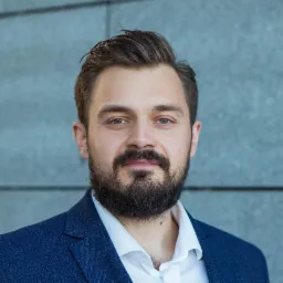 Portret brodatego mężczyzny w niebieskiej marynarce i białej koszuli na tle szarej ściany z wyraźną fakturą.