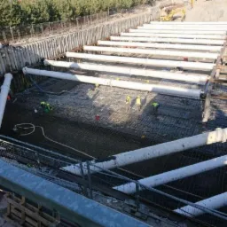 Głęboki wykop zabezpieczony rurami i ściankami szczelinowymi, widoczna siatka zbrojeniowa i pracownicy w kamizelkach odblaskowych przygotowujący fundamenty, w tle koparka i pryzma piasku.