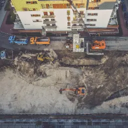 Zdjęcie z lotu ptaka przedstawia plac budowy z dwoma koparkami, dźwigiem, kontenerami, dwiema ciężarówkami i częściowo wzniesionym budynkiem mieszkalnym w tle.