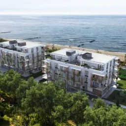 Wizualizacja dwóch nowoczesnych, wielopiętrowych apartamentowców z płaskimi dachami, z widokiem na morze i plażę, otoczonych zielenią drzew i krzewów.