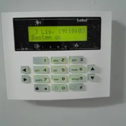 Biały panel sterowania systemu alarmowego Satel CA-10 z wyświetlaczem LCD wskazującym datę, godzinę i status 'System ok', przyciski numeryczne i funkcyjne z podświetleniem LED.