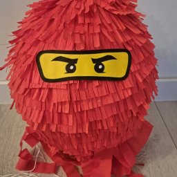 ANIMATOR Izabela Zbiega - Czerwona piniata w kształcie kuli z papierowymi frędzlami, stylizowana na głowę postaci z Lego Ninjago, z żółtymi oczami na czarnym tle.