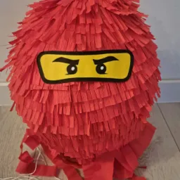 Czerwona piniata w kształcie kuli z papierowymi frędzlami, stylizowana na głowę postaci z Lego Ninjago, z żółtymi oczami na czarnym tle.