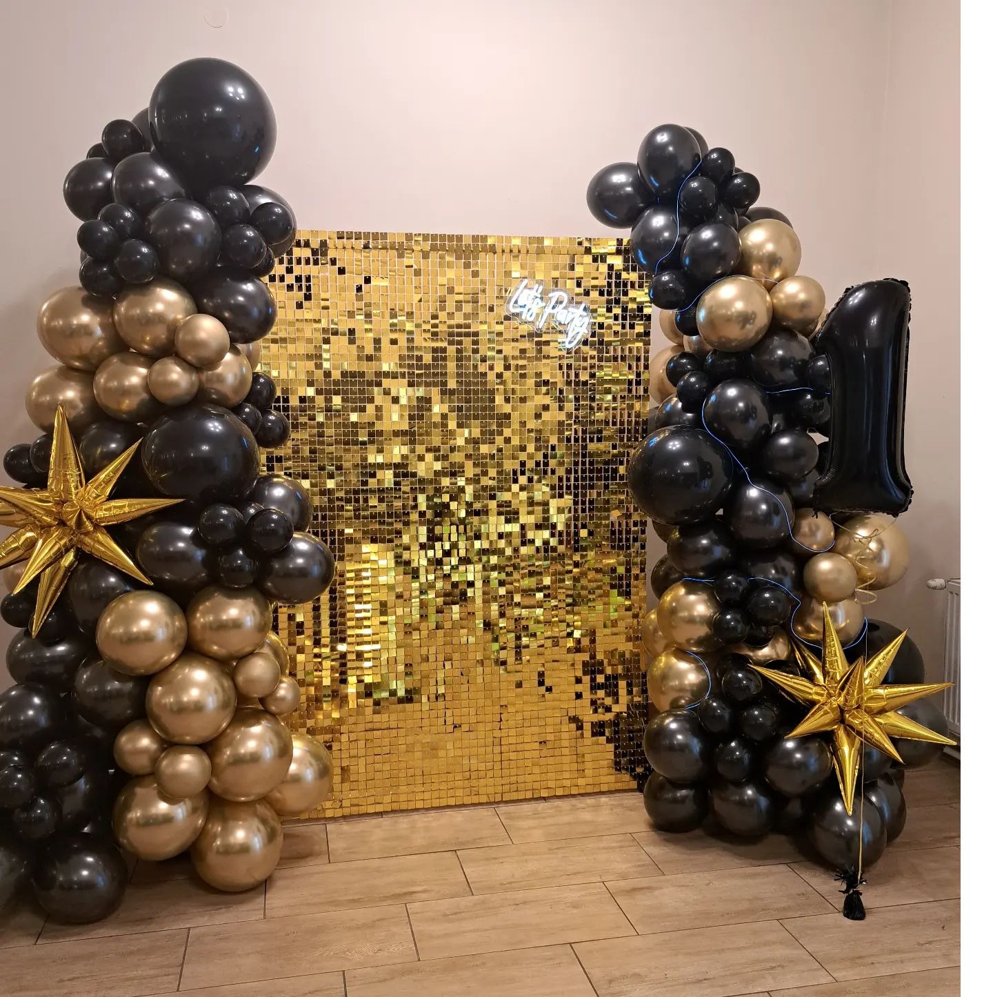 Dekoracja urodzinowa: ścianka z błyszczących złotych kwadratów z neonowym napisem 'Let's Party', otoczona girlandami z czarnych i złotych balonów, z dodatkowymi złotymi gwiazdami i balonem...