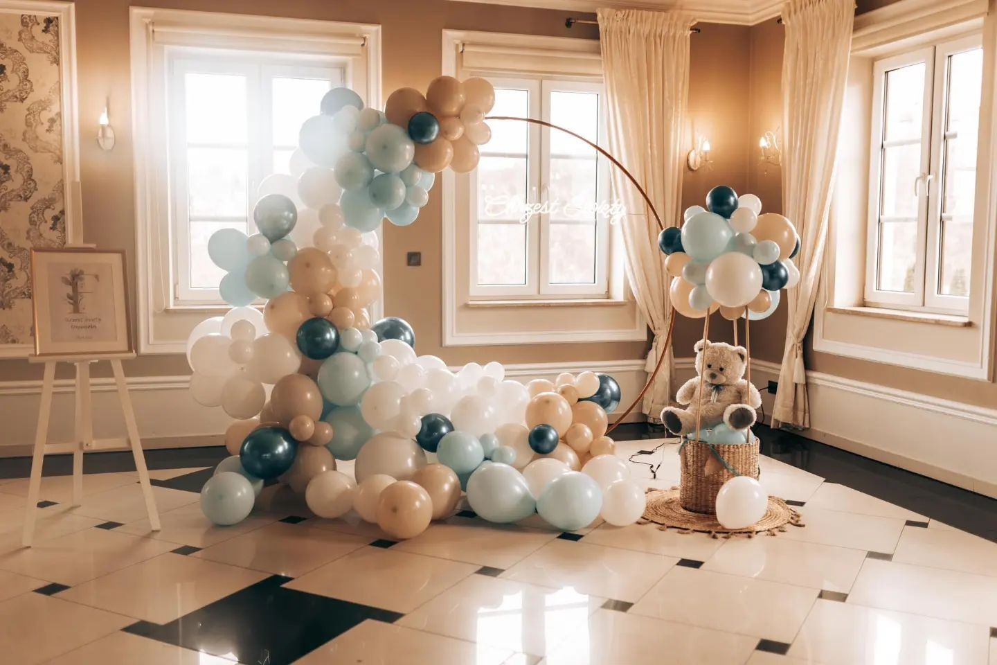 Dekoracja balonowa w odcieniach bieli, beżu i błękitu, ułożona w kształt półksiężyca z dodatkiem pluszowego misia w koszu, całość na tle eleganckiego wnętrza z dużymi oknami.