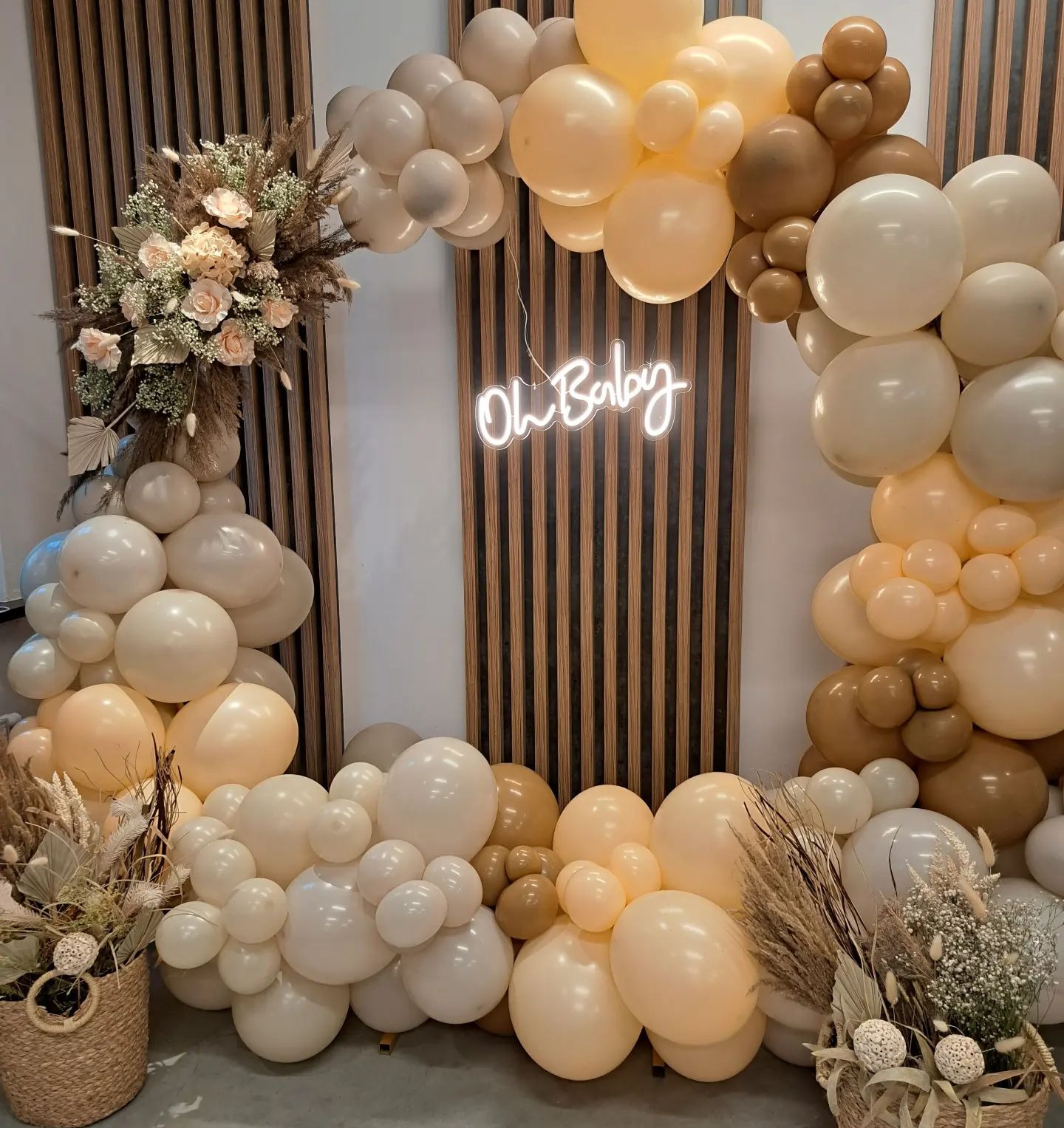 Dekoracja z beżowych i białych balonów z neonowym napisem 'Oh Baby' na tle drewnianej ścianki, uzupełniona kompozycjami kwiatowymi w koszykach.