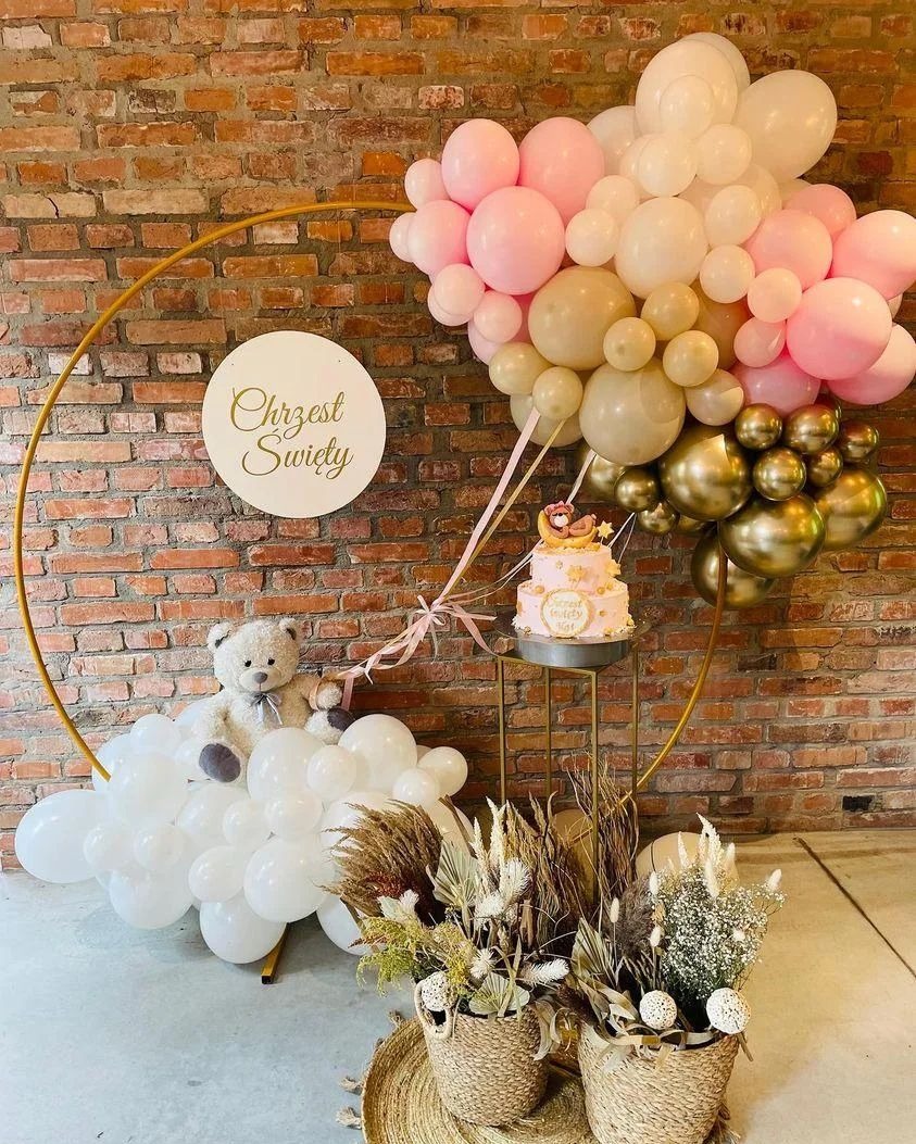 Dekoracja na chrzest święty z tortem, balonami w kolorach pastelowych, złotym okręgiem i pluszowym misiem na tle ceglanej ściany.