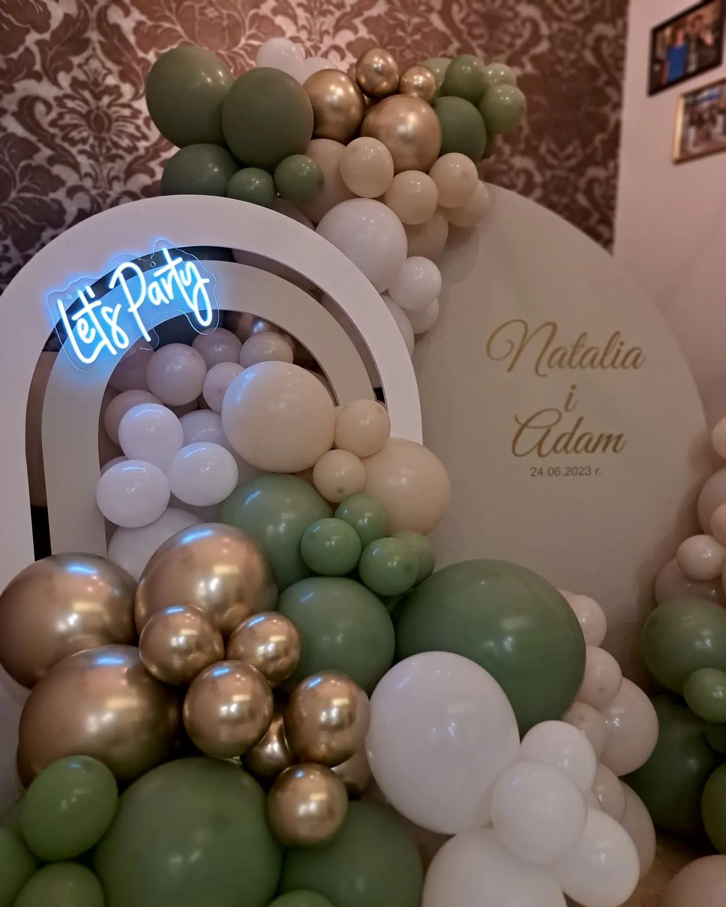 Dekoracja balonowa w odcieniach bieli, złota i zieleni zdobiąca biały łuk z napisem neonowym 'Let's Party' oraz ściankę z imionami 'Natalia i Adam' i datą 24.06.2023, wykonana w Bochni.