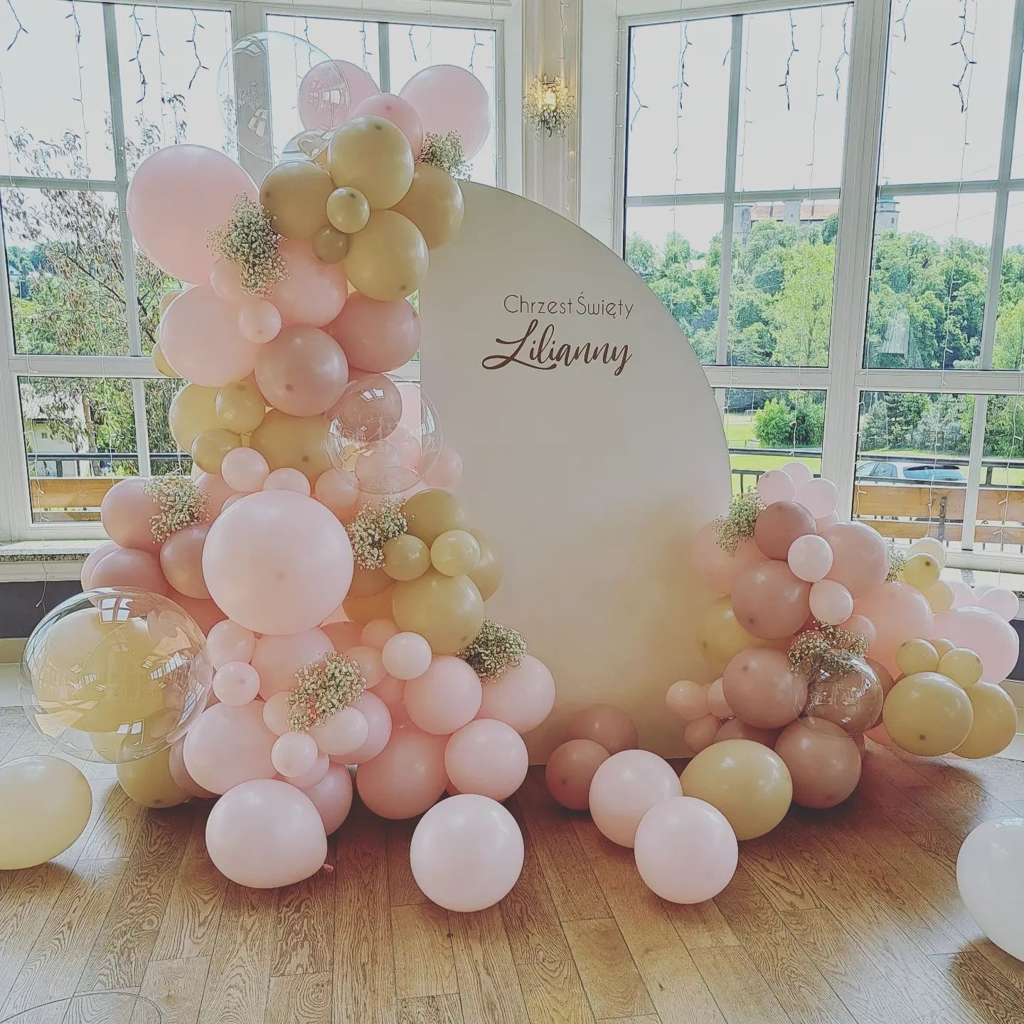 Dekoracja balonowa w odcieniach różu i beżu z białym półokręgiem z napisem 'Chrzest Święty Lilianny', ustawiona na drewnianej podłodze przed oknami z widokiem na zieleń.