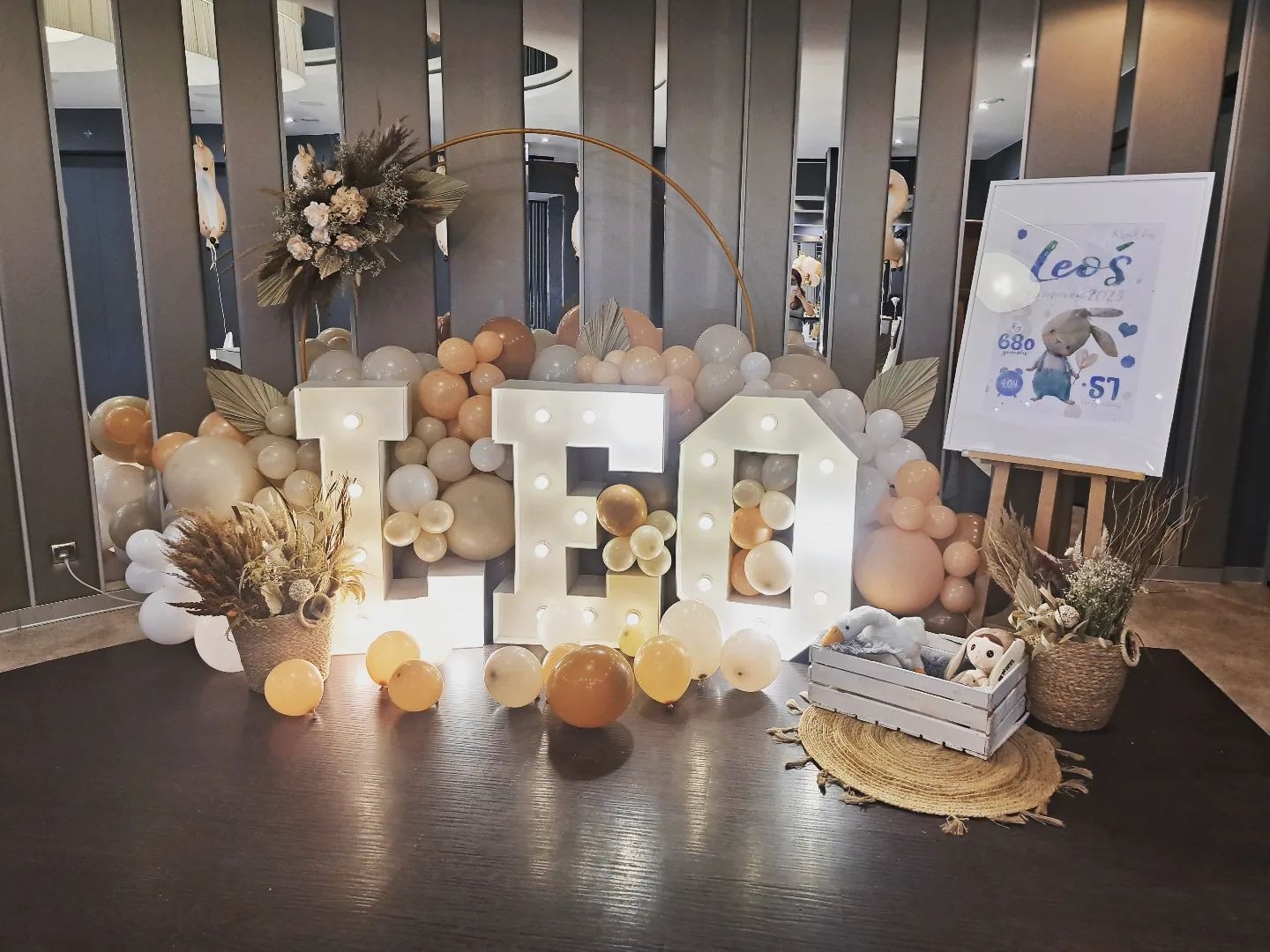 Dekoracja urodzinowa z balonów w odcieniach beżu i bieli, podświetlane litery 'LEO', plakat z motywem króliczka na sztaludze, kompozycje z suszonych kwiatów w koszykach i pluszowe zabawki w białej...