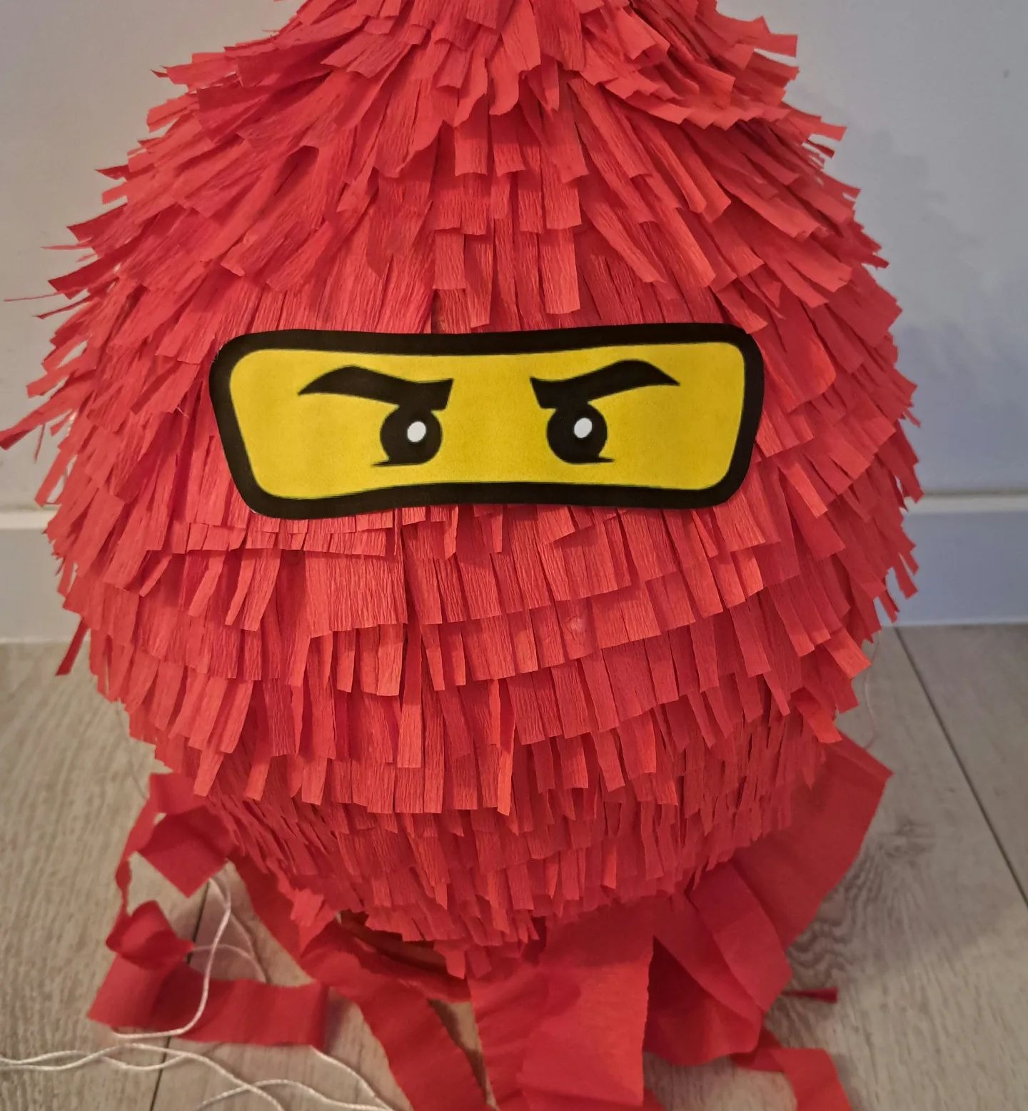 Czerwona piniata w kształcie kuli z papierowymi frędzlami, stylizowana na głowę postaci z Lego Ninjago, z żółtymi oczami na czarnym tle.