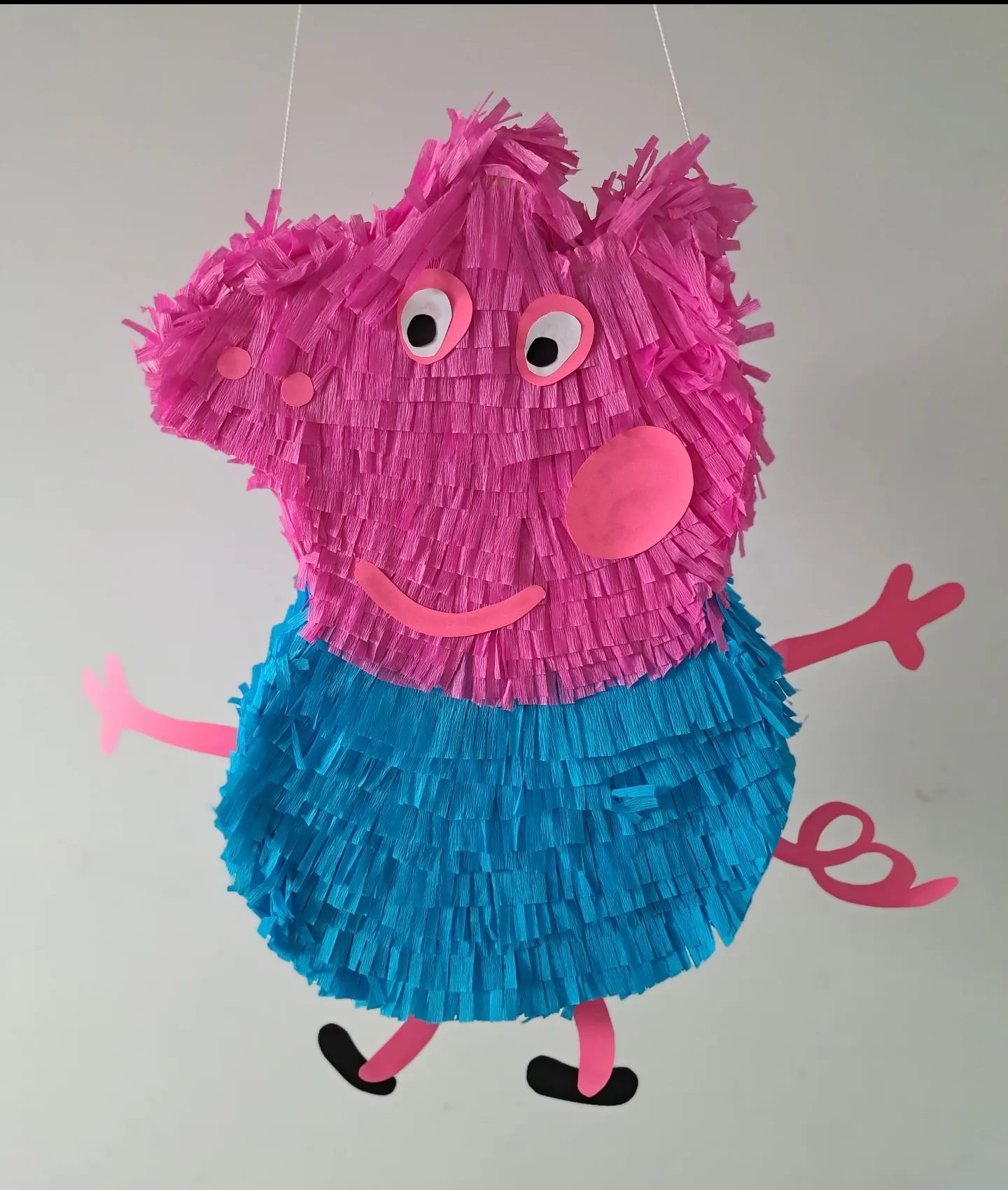 Ręcznie robiona piniata w kształcie świnki Peppy, wykonana z różowej i niebieskiej krepy, zawieszona na białym sznurku.