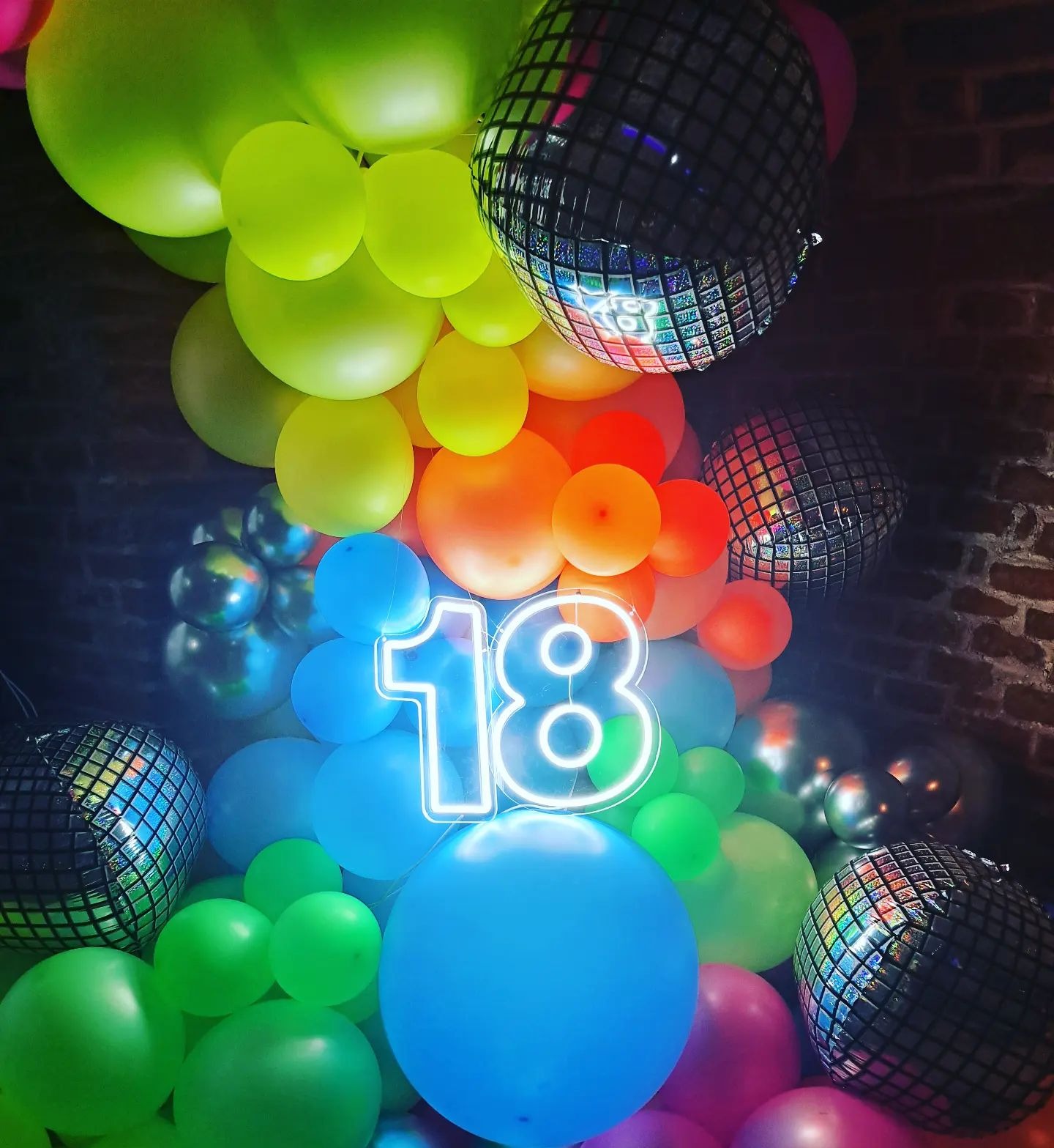 Kolorowa dekoracja urodzinowa z balonów w odcieniach tęczy, lustrzanymi kulami dyskotekowymi i neonowym napisem '18' na tle ceglanej ściany.
