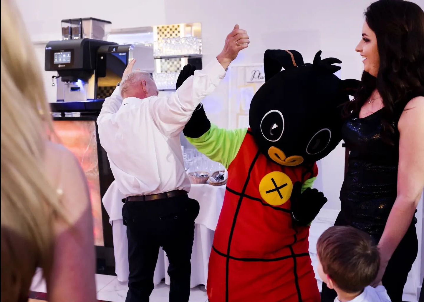 Osoba w kostiumie Binga tańczy z gośćmi na przyjęciu, widoczna ekspresowa maszyna do kawy w tle.