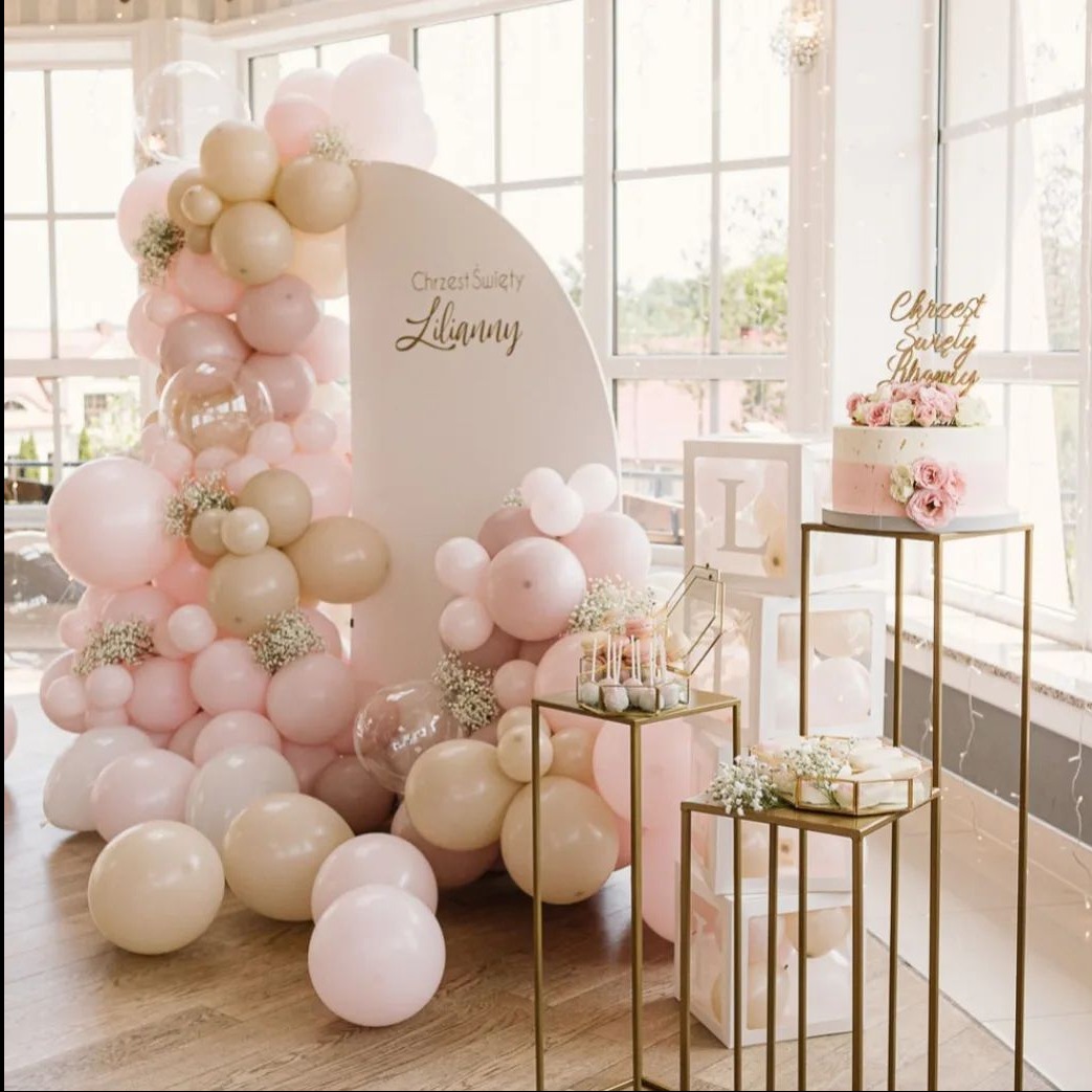 Dekoracja balonowa w odcieniach różu i beżu z białym tłem z napisem 'Chrzest Święty Lilianny', tort z kwiatami na złotym stojaku oraz dekoracyjne pudełka z literą 'L', ustawione na tle okna.