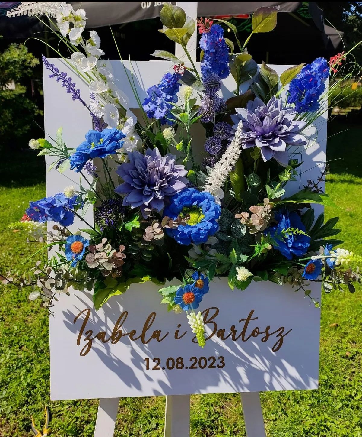 Dekoracja kwiatowa na białej tablicy z imionami Izabela i Bartosz oraz datą 12.08.2023, ustawiona na trawniku w plenerze.
