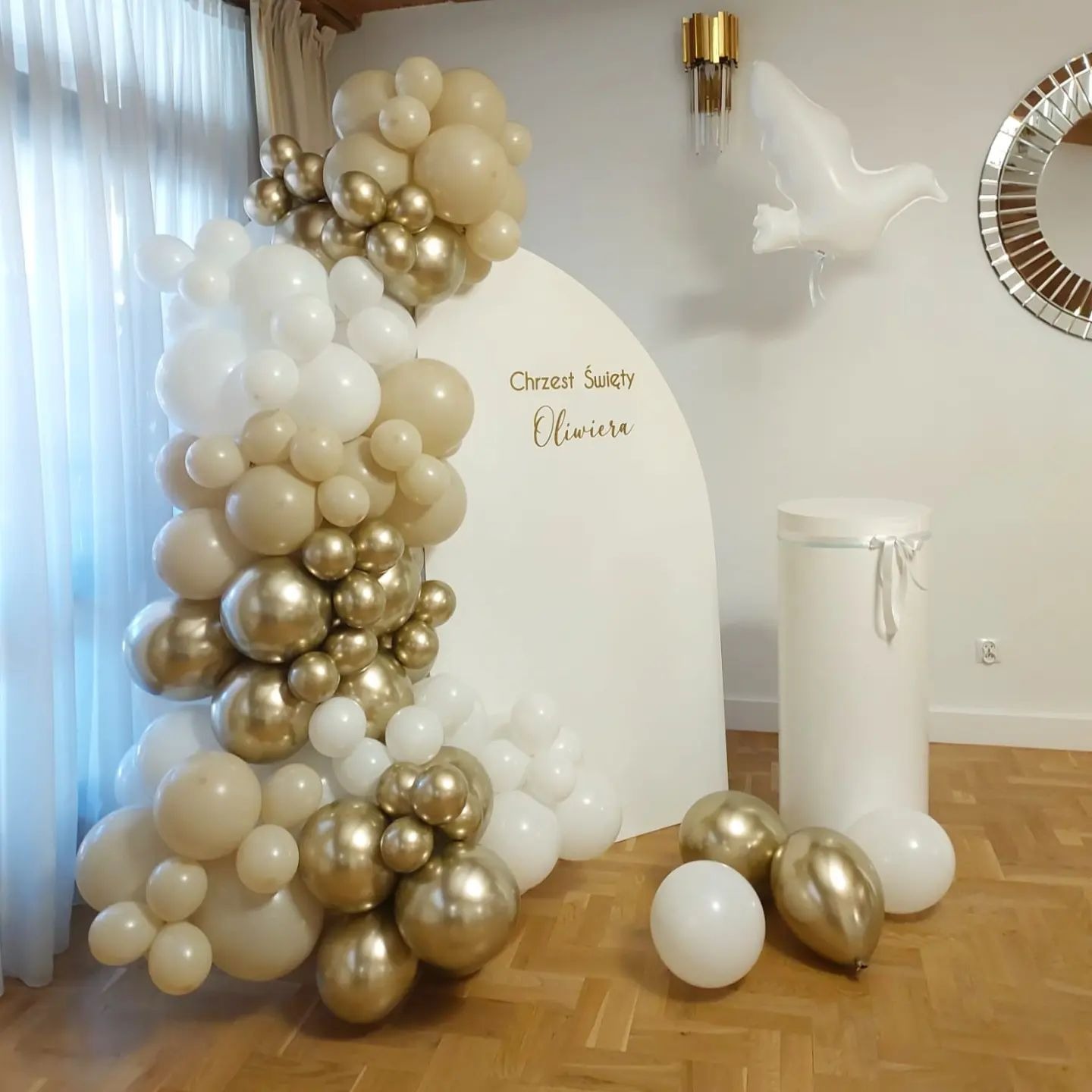 Dekoracja balonowa w odcieniach bieli, beżu i złota z napisem 'Chrzest Święty Oliwiera' na białym tle, uzupełniona o balon w kształcie gołębia i białą kolumnę, tworząca elegancką aranżację...