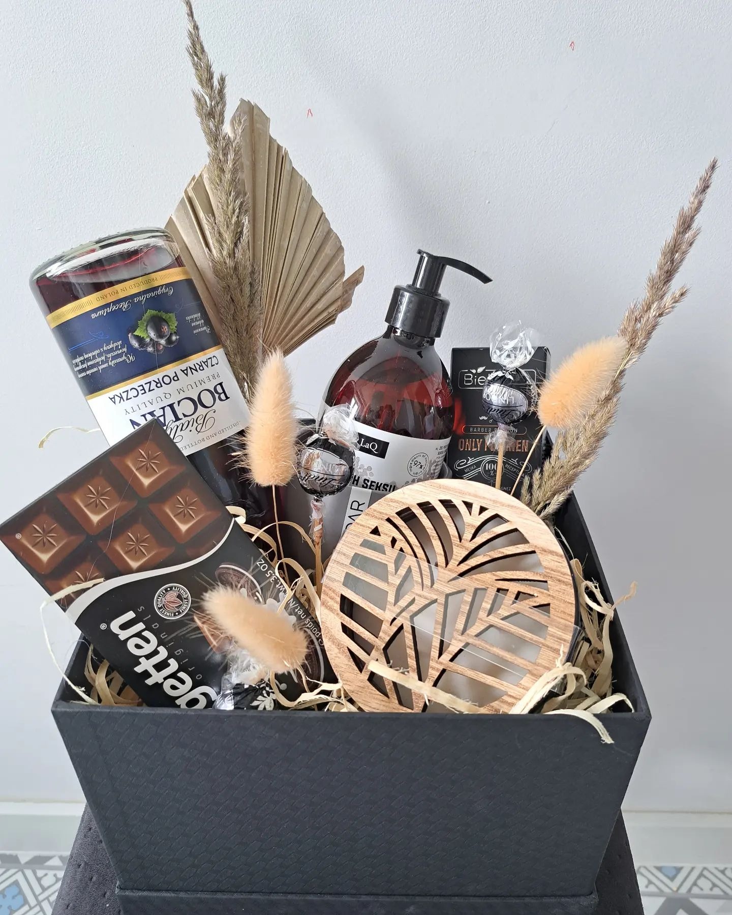 Elegancki, czarny kosz prezentowy wypełniony butelką nalewki z czarnej porzeczki, czekoladą Getten, kosmetykami LaQ i Bielszy, oraz dekoracyjnym drewnianym podkładką z motywem liści, całość...