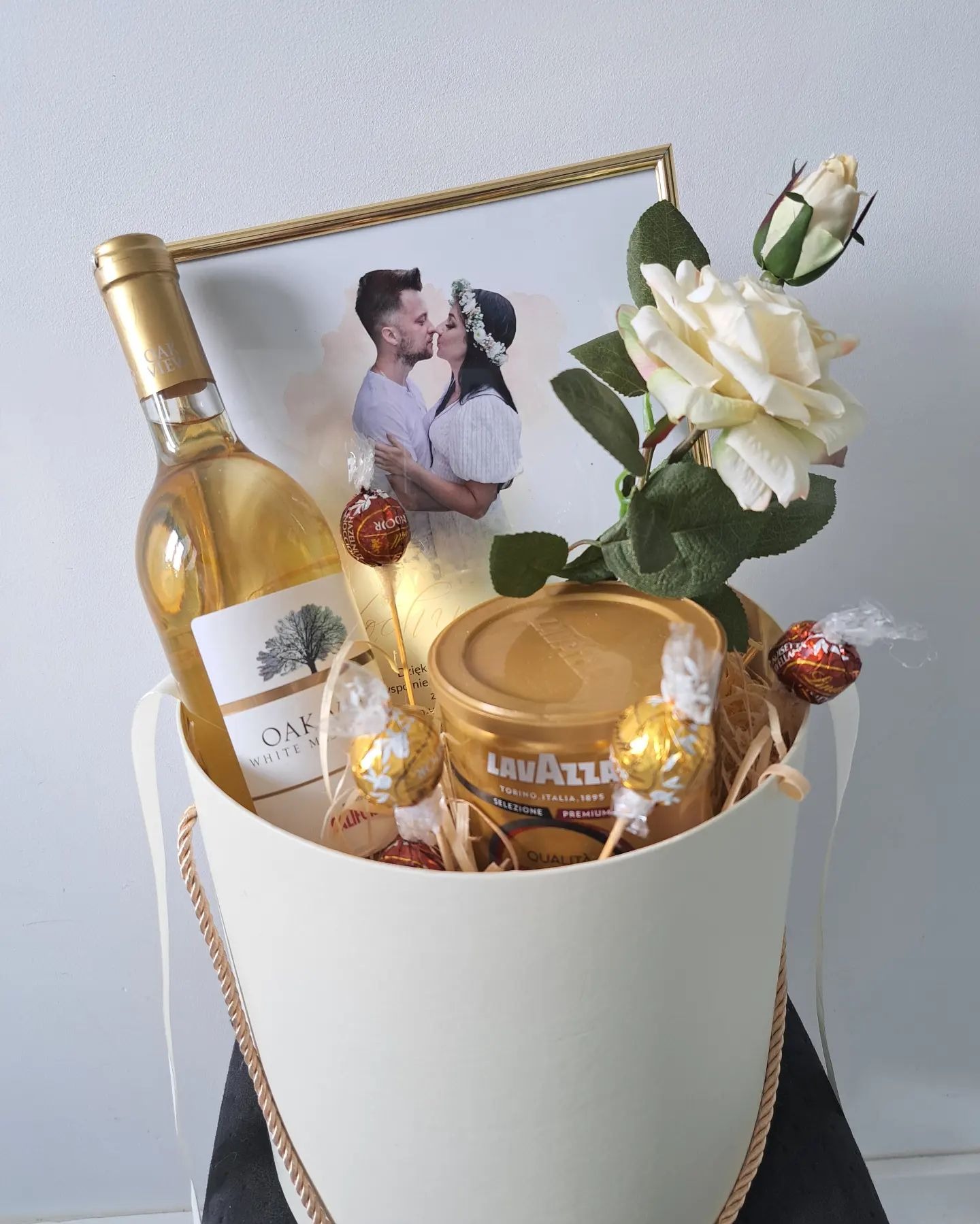Kosz prezentowy z winem Oak White, kawą Lavazza, czekoladkami Ferrero Rocher, sztuczną białą różą i oprawionym zdjęciem całującej się pary.
