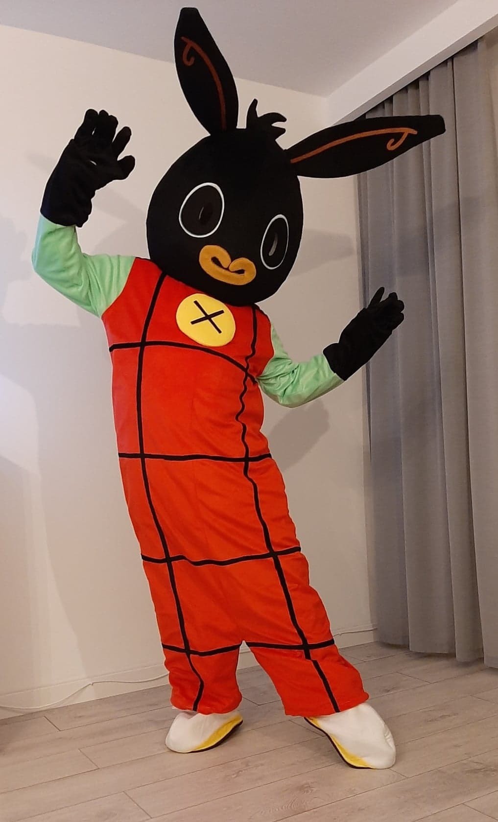 Osoba w kostiumie królika Binga, z uniesionymi rękami, na jasnym tle ściany i zasłony.
