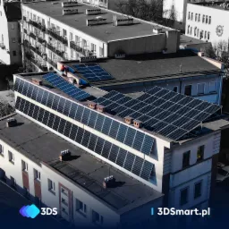 Widok z góry na budynek z zamontowanymi panelami słonecznymi na dachu, instalator w czerwonym kasku na dachu, częściowo odbarwione tło.