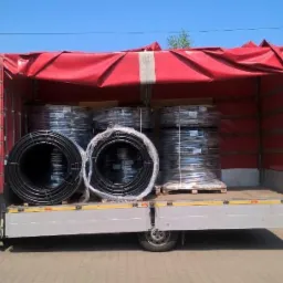 Biały bus z otwartymi drzwiami, załadowany rolkami czarnych rur w przezroczystej folii, pod czerwonym brezentem, na tle budynku gospodarczego i drabiny.