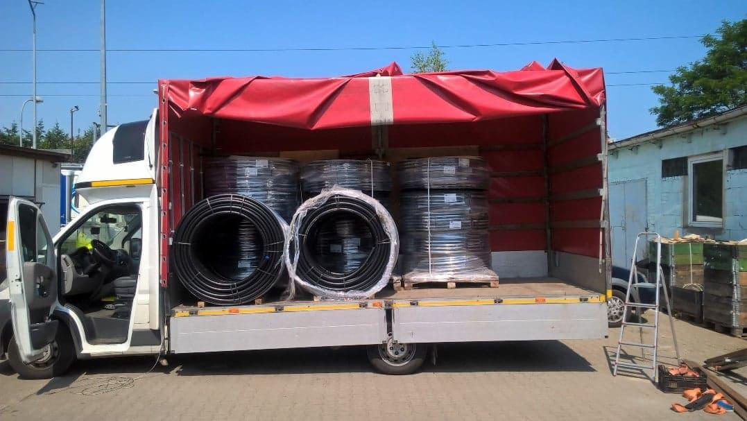 Biały bus z otwartymi drzwiami, załadowany rolkami czarnych rur w przezroczystej folii, pod czerwonym brezentem, na tle budynku gospodarczego i drabiny.