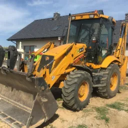 Żółta koparko-ładowarka JCB 3CX na placu budowy, z widocznym wykopem i fragmentem nowo wybudowanego domu w tle, w słoneczny dzień.