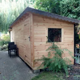 Drewniana konstrukcja z oknem, prawdopodobnie domek narzędziowy, z ciemnym dachem z papy i dwoma krzesłami obok, na tle zieleni ogrodu.