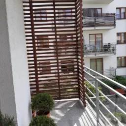 Balkon z drewnianą ażurową przesłoną zapewniającą prywatność, widok z boku, rośliny w donicach na pierwszym planie, balustrada ze szkła i stali nierdzewnej, cień rzucany przez przesłonę na podłogę...