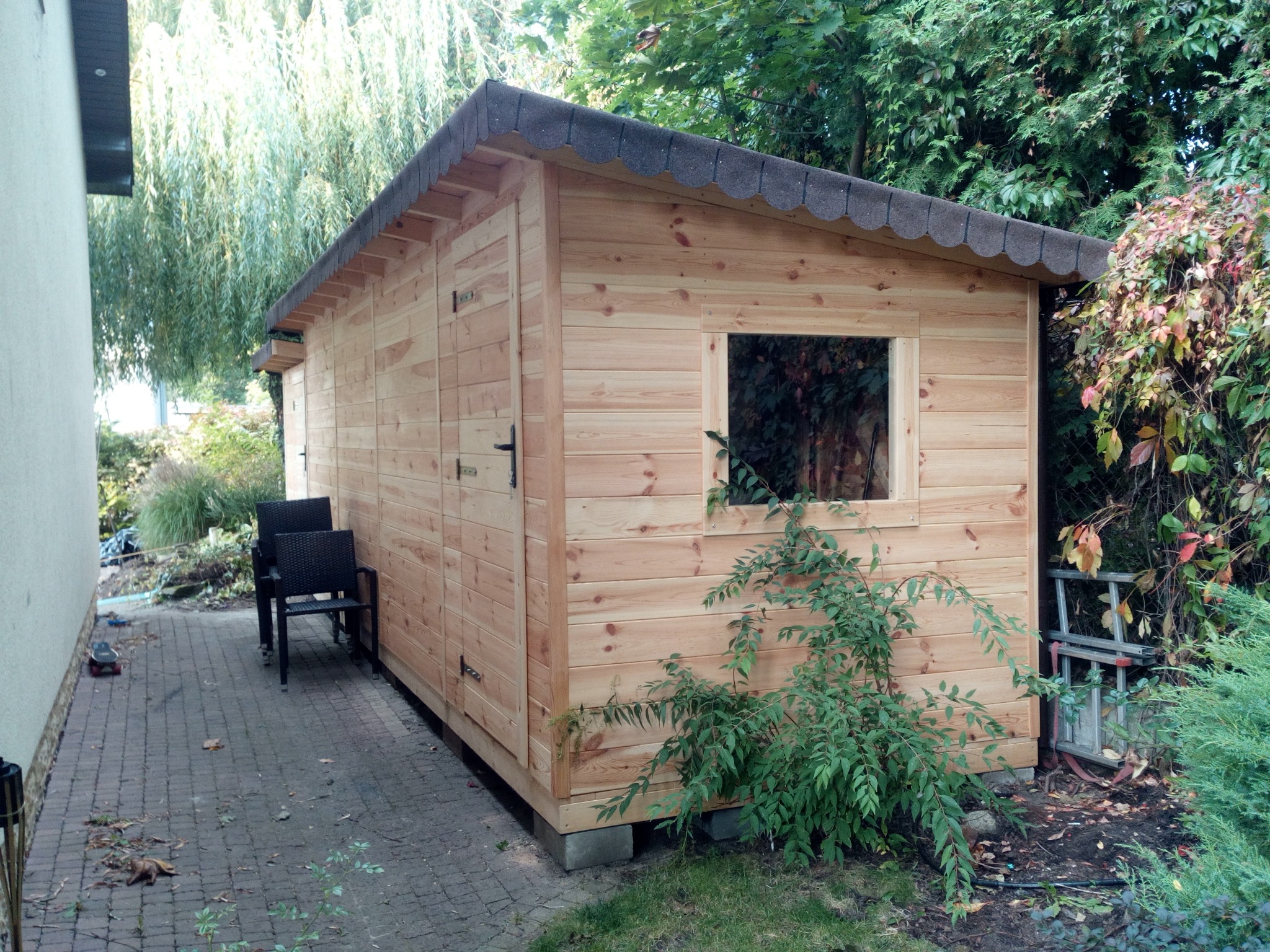 Drewniana konstrukcja z oknem, prawdopodobnie domek narzędziowy, z ciemnym dachem z papy i dwoma krzesłami obok, na tle zieleni ogrodu.