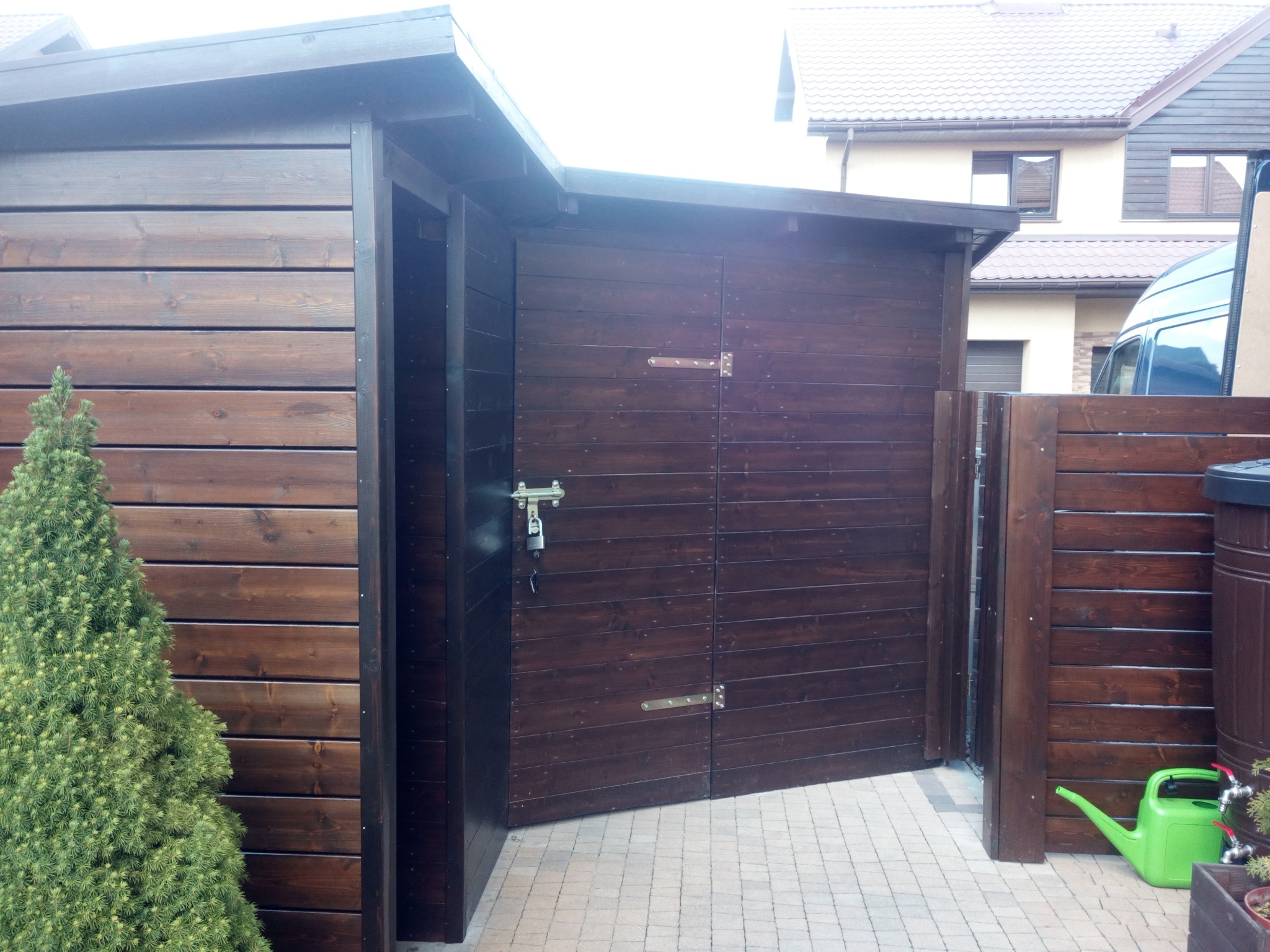 Ciemnobrązowa, drewniana konstrukcja z podwójnymi drzwiami, zamkniętymi na kłódkę, z widoczną zieloną konewką i małym drzewkiem iglastym obok, na tle kostki brukowej i fragmentu domu.