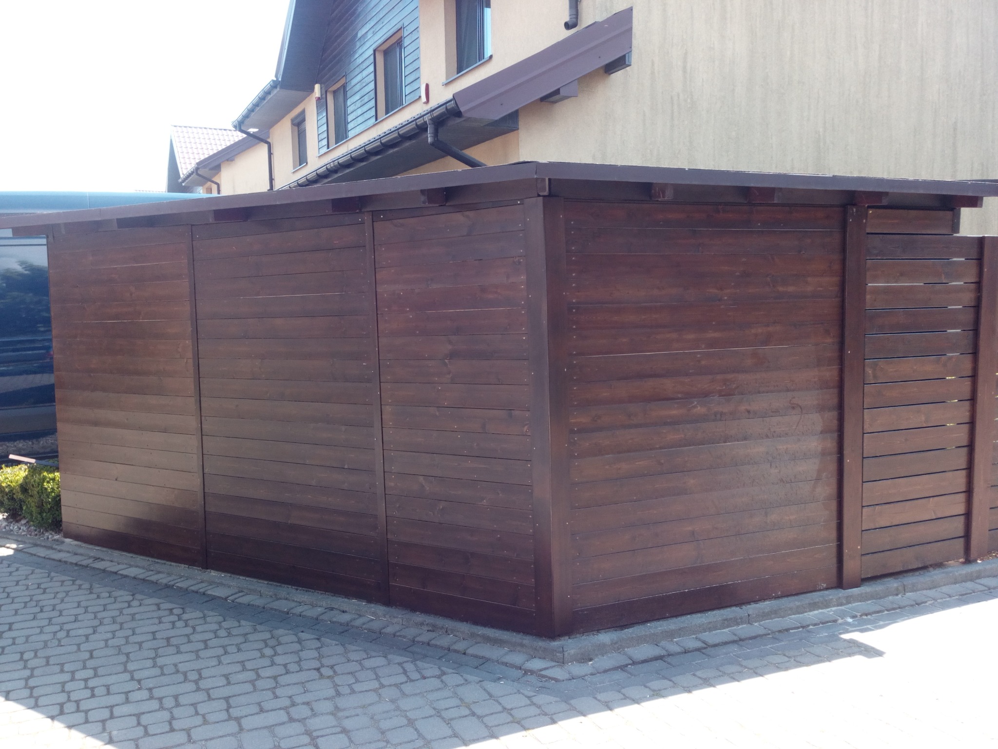 Ciemnobrązowa, drewniana konstrukcja z poziomych desek, prawdopodobnie szopa lub składzik ogrodowy, przylegająca do budynku, z kostką brukową na pierwszym planie.