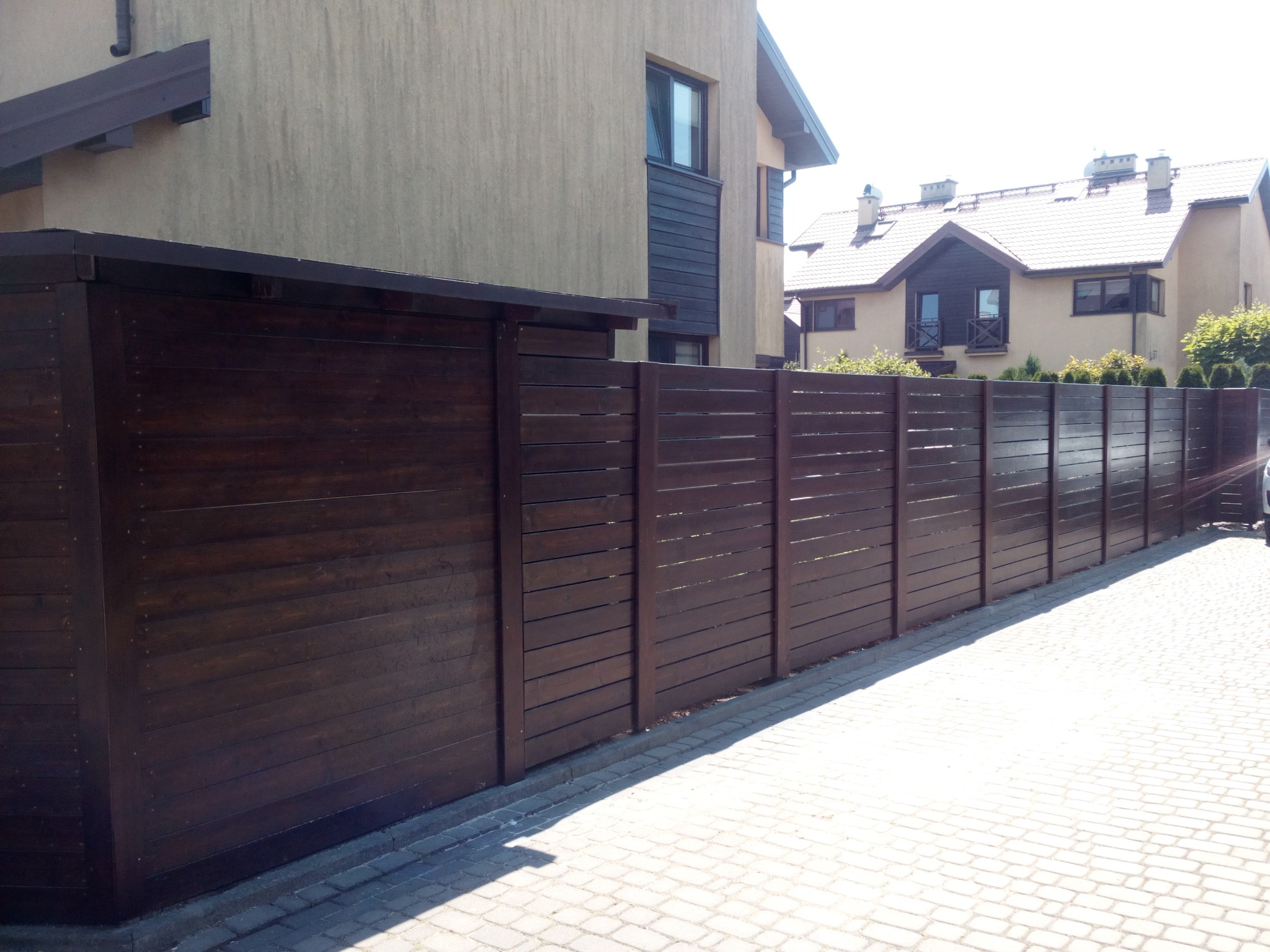 Ciemnobrązowe, poziome deski tworzące nowoczesne ogrodzenie posesji z widocznymi domami w tle, oświetlone jasnym światłem słonecznym.