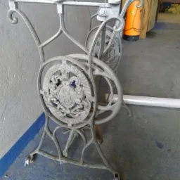 Stelaż stołu z żeliwa z dekoracyjnym motywem herbowym, pomalowany na szaro, z widocznymi śladami użytkowania i kółkami u podstawy, na tle szarej ściany i betonowej podłogi.