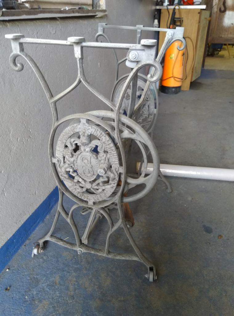 Stelaż stołu z żeliwa z dekoracyjnym motywem herbowym, pomalowany na szaro, z widocznymi śladami użytkowania i kółkami u podstawy, na tle szarej ściany i betonowej podłogi.