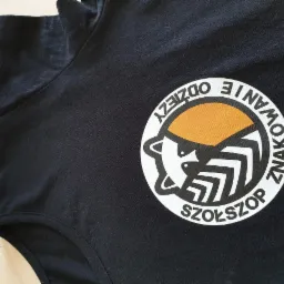 Czarna koszulka z okrągłym nadrukiem logo firmy 'SZOLSZOP' z elementami graficznymi w kolorze białym i pomarańczowym, leżąca na drewnianym tle.