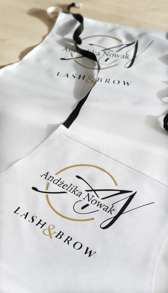 Dwa białe fartuchy z nadrukowanym logo 'Andżelika Nowak LASH&BROW' z czarnymi wiązaniami, leżące na jasnym tle.