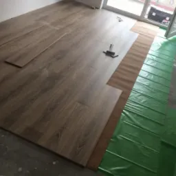 Układanie paneli podłogowych w pokoju, widoczny młotek, podkład korkowy i zielona folia izolacyjna na surowym betonie.