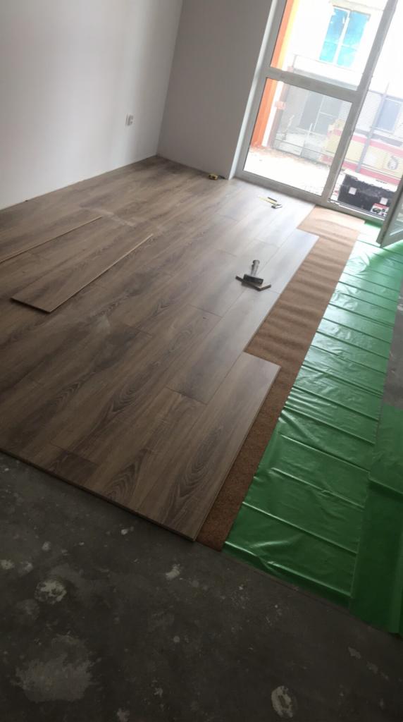 Układanie paneli podłogowych w pokoju, widoczny młotek, podkład korkowy i zielona folia izolacyjna na surowym betonie.