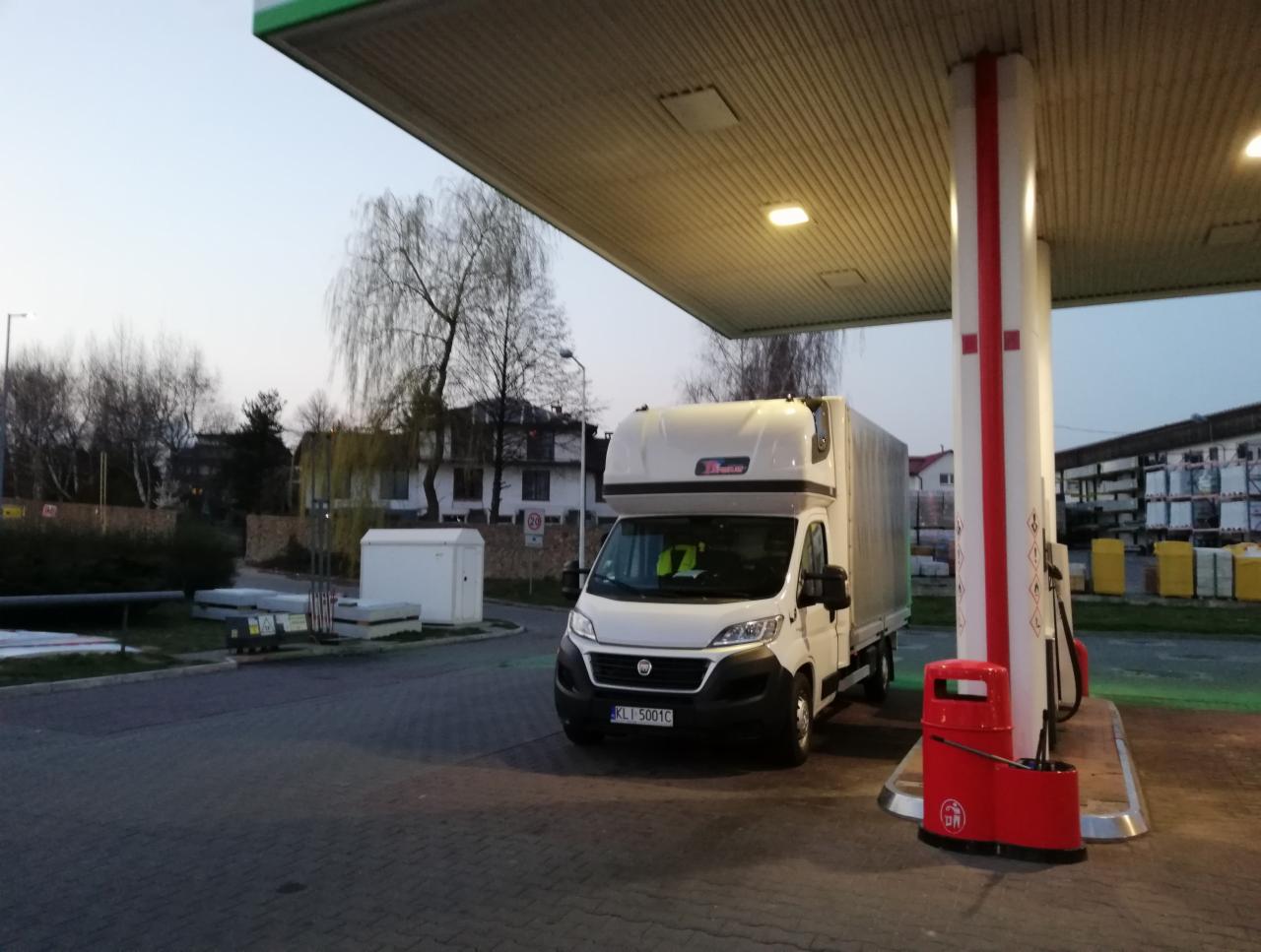 Biały samochód dostawczy typu furgon z plandeką na stacji benzynowej o zmierzchu, widoczny numer rejestracyjny i odblaskowa kamizelka kierowcy.