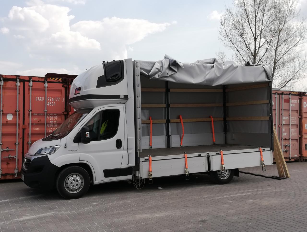 Biała ciężarówka z otwartą przestrzenią ładunkową, zabezpieczona szarym plandekowym dachem i pomarańczowymi pasami mocującymi, zaparkowana na placu z widocznymi czerwonymi kontenerami w tle.