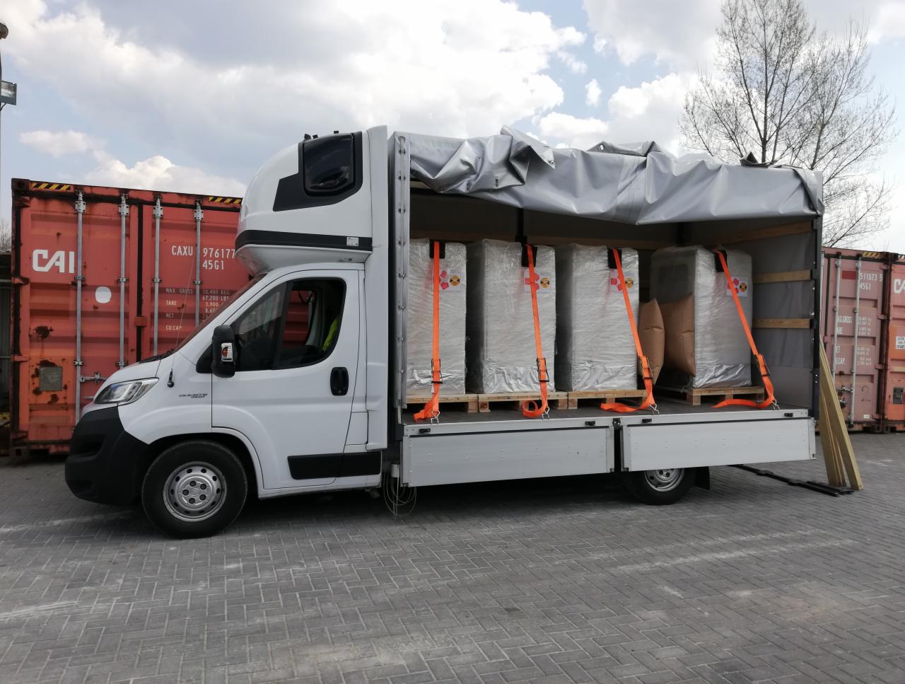 Biała furgonetka z otwartą przestrzenią ładunkową załadowana paletami z towarem owiniętym folią i zabezpieczonym pomarańczowymi pasami transportowymi, na tle czerwonych kontenerów.