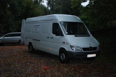 Biały samochód dostawczy Mercedes Sprinter z widoczną tablicą rejestracyjną na tle leśnego parkingu z opadłymi liśćmi, zaparkowany obok srebrnego kombi.