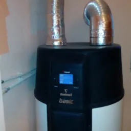 Pionowy piec grzewczy Galmet Basic z dwoma elastycznymi przewodami wentylacyjnymi przymocowanymi u góry, zainstalowany w rogu pomieszczenia z widocznymi rurami instalacji.